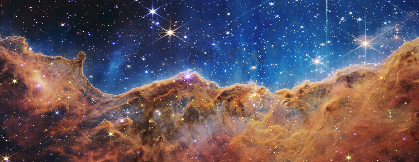 JWST image of the Cosmic Cliffs showing a long jagged ridge of glowing orange-brown gas and dust below a blue star-filled background, with many bright young stars embedded around the ridge.