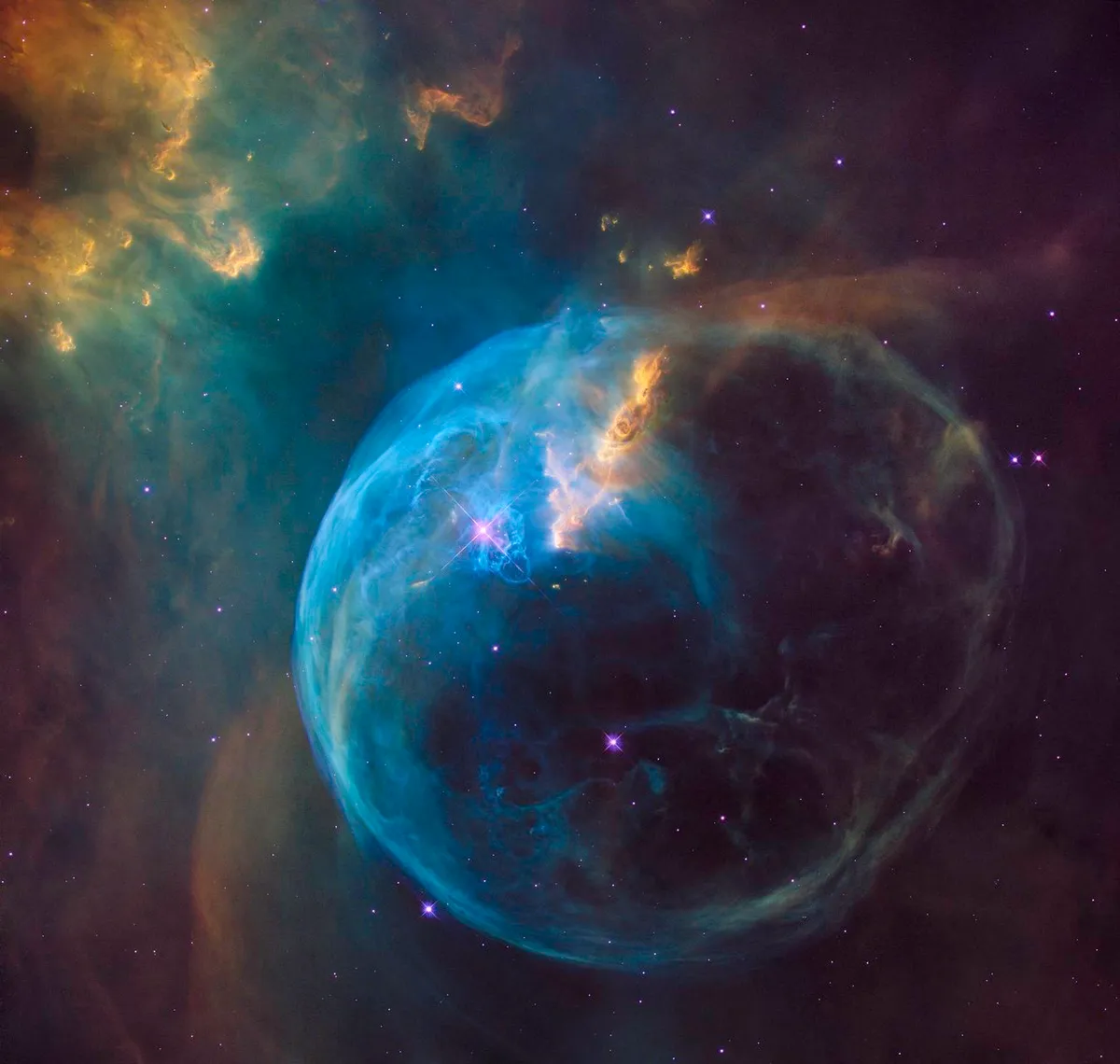 Color image of the Bubble Nebula showing a large blue spherical shell of glowing gas surrounded by darker space and warmer orange clouds, with bright stars embedded nearby.