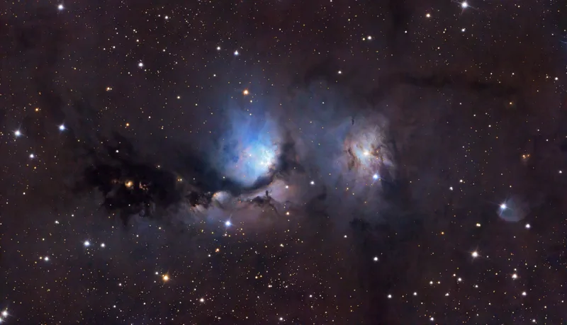 Wide-field image of the reflection nebula M78 showing two bright blue glowing patches around embedded stars, crossed by dark dusty lanes against a star-filled background.