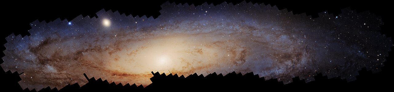 Long panoramic Hubble image of the Andromeda galaxy showing a bright yellow-white bulge, blue star-forming disk regions, dust lanes, and a wide field of resolved stars.