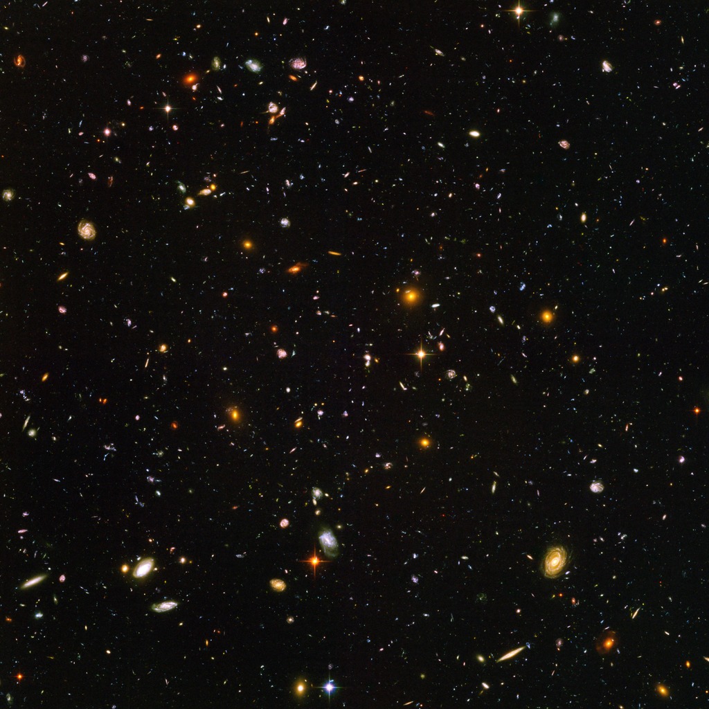Hubble Ultra Deep Field image showing a dark patch of sky filled with hundreds of tiny galaxies of many colors and shapes, plus a few foreground stars.