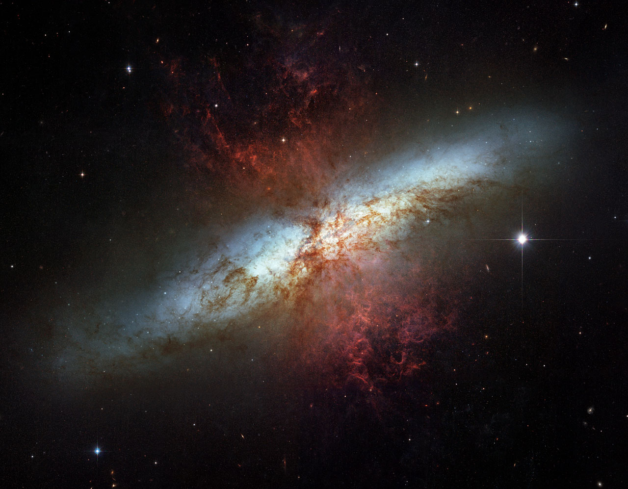 Image of the starburst galaxy M82 showing a bright blue-white disk crossed by dust lanes and red glowing gas streaming out above and below the galaxy.