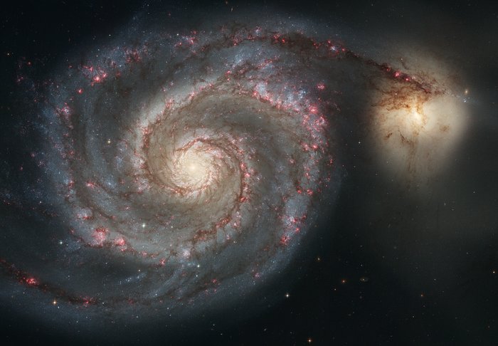 Hubble image of the Whirlpool Galaxy with a bright spiral disk, pink star-forming regions, dark dust lanes, and a smaller companion galaxy connected by tidal structure.