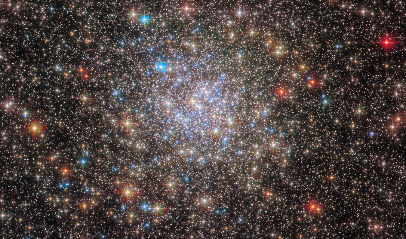 Dense star field image with a bright cluster near the center and many stars of different apparent colors, including blue-white, yellow, and red-orange stars scattered across the frame.