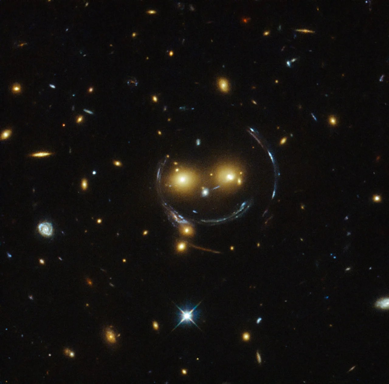 Hubble image of a galaxy cluster with several bright foreground galaxies and curved blue arcs produced by strong gravitational lensing of background galaxies.