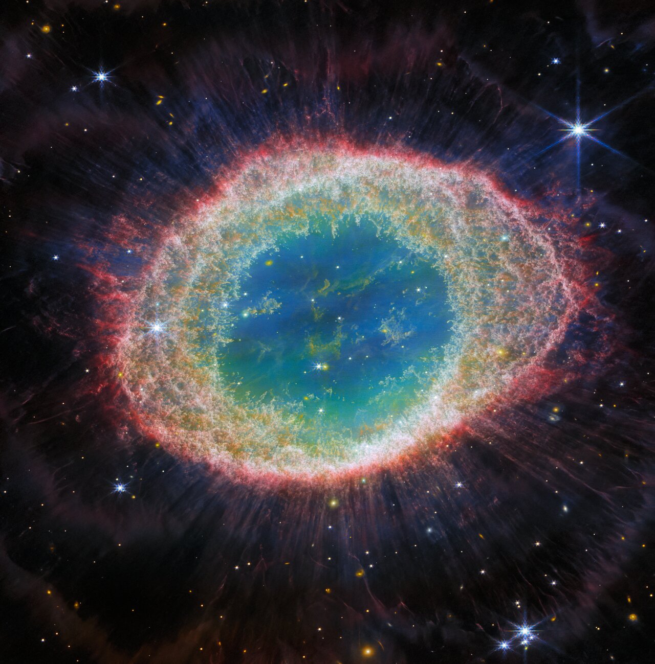 JWST image of the Ring Nebula showing a bright elliptical ring of glowing gas with intricate filamentary structure surrounding a small hot central star. The nebula is set against a dark background with many faint stars.