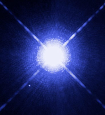 Hubble Space Telescope image of the Sirius binary system. Sirius A is a brilliant overexposed white star dominating the frame with prominent diffraction spikes. Sirius B is visible as a small faint dot at lower left, separated by several arcseconds.